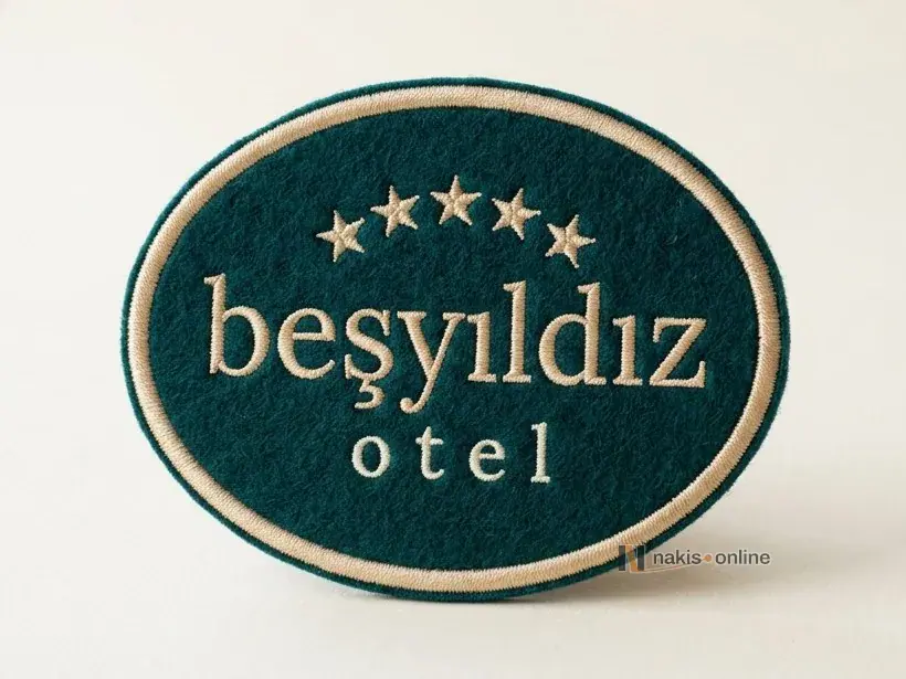 Açık renk stüdyo zemininde koyu zümrüt yeşili mat çuha zeminli, şampanya altını saten kenarlı oval blazer arması; kavis çizen beş yıldız ve altında bir otelin tırnaklı harfli adı