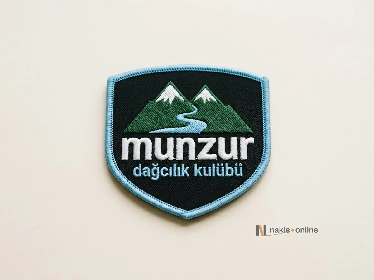 Açık renk stüdyo zemininde siyah dimi zeminli, buz mavisi merrow kenarlı kalkan biçimli kulüp arması; karlı ikiz zirve simgesi ve beyaz harflerle bir dağcılık kulübünün adı