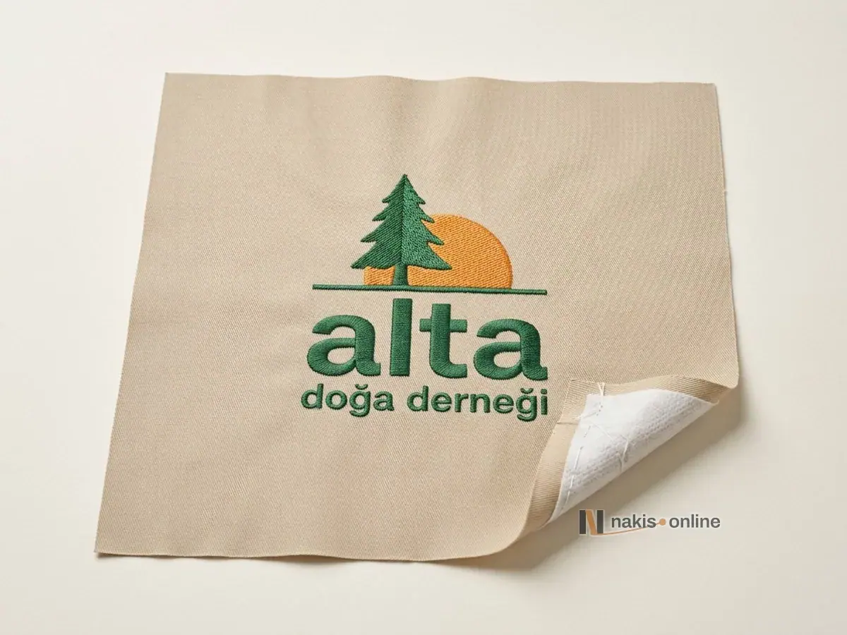 Açık renk stüdyo zemininde kum bej dimi deneme parçası; ortasında çam ağacı ve yarım güneş simgeli bir doğa derneğinin logosu saten ve dolgu dikişle işlenmiş, kıvrık köşesinde beyaz yırtma tela ve alt iplik görünüyor
