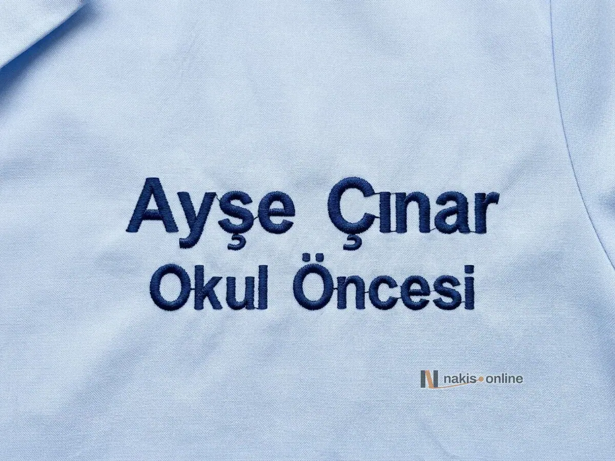 Açık mavi öğretmen önlüğünün göğsünde yakın çekim isim nakışı; lacivert harflerle iki satır, ad soyad ve branş, Türkçe karakter işaretleri seçiliyor