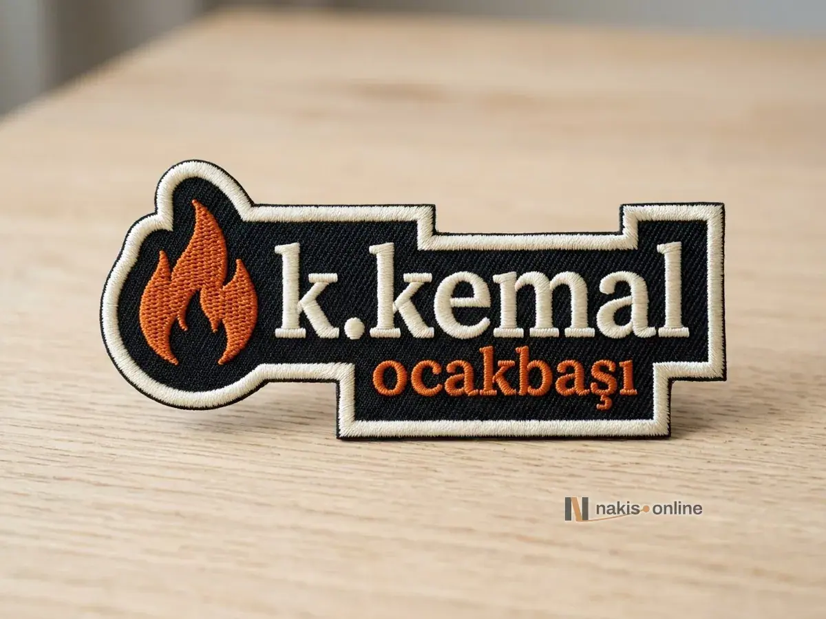 Logo hattından kesilmiş siyah dimi armanın yakın çekimi; turuncu alev simgesi, krem tırnaklı harflerle bir ocakbaşı restoranının adı ve girintili formu saran krem saten kenar