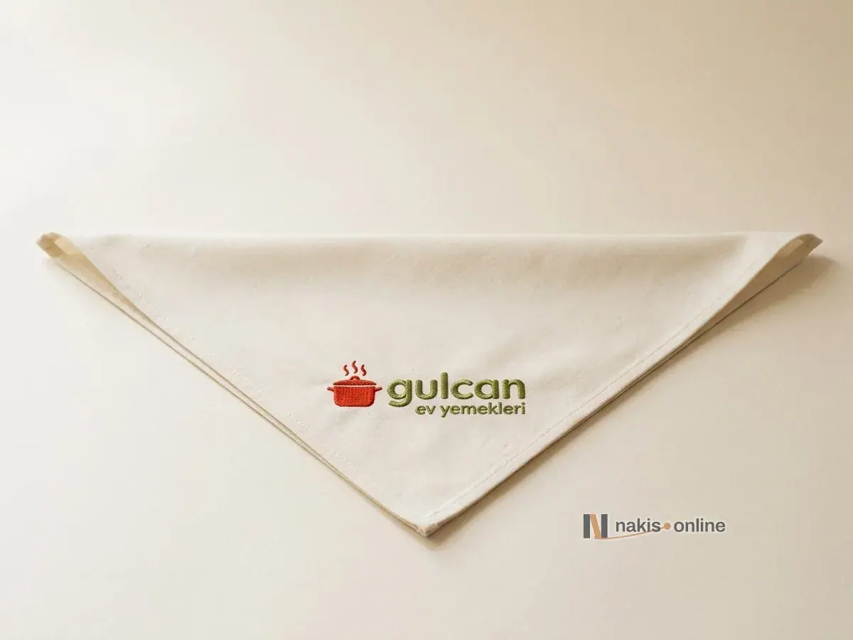 Stüdyo zemininde üçgen katlanmış ekru pamuk bandana; ucuna yakın, bağlanınca düz okunacak yönde domates kırmızısı tencere simgeli, zeytin yeşili harfli bir ev yemekleri lokantasının logosu