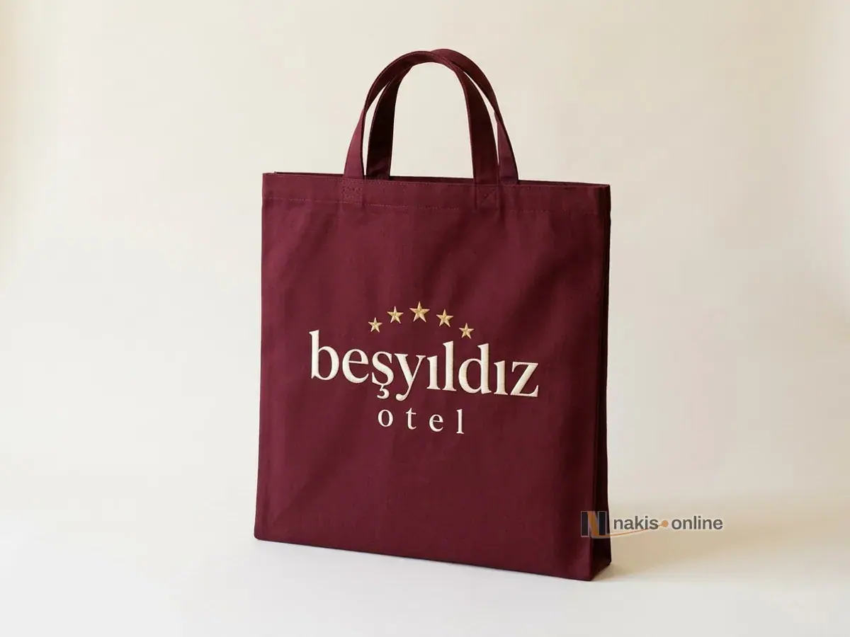 Bordo bez çanta: ön yüzde kemer biçiminde dizilmiş altın sarısı yıldızlar ve krem iplikle işlenmiş tırnaklı harflerle bir otelin logosu