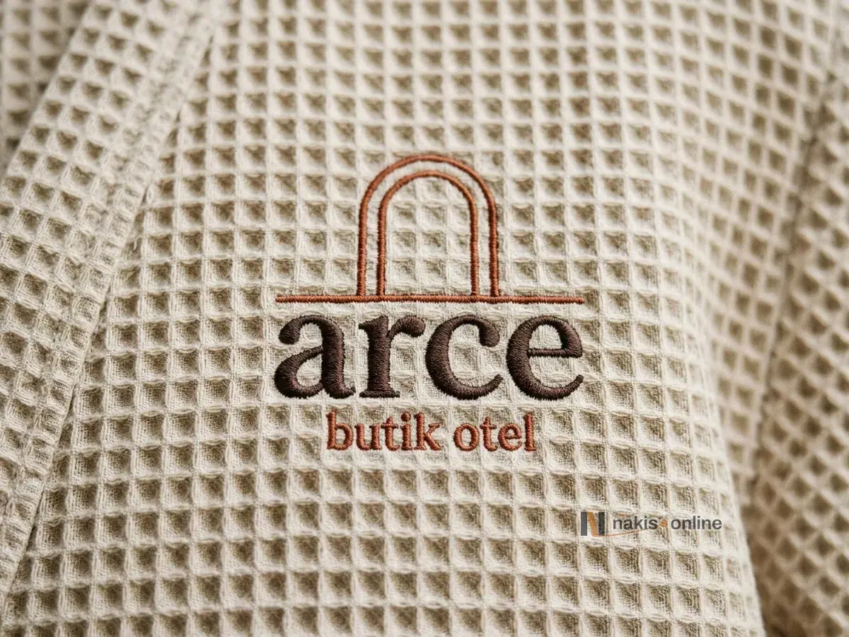 Bej petek dokulu bornozun göğsünde kiremit rengi kemerli kapı simgesi ve koyu kahve harflerle işlenmiş bir butik otel logosunun makro görüntüsü