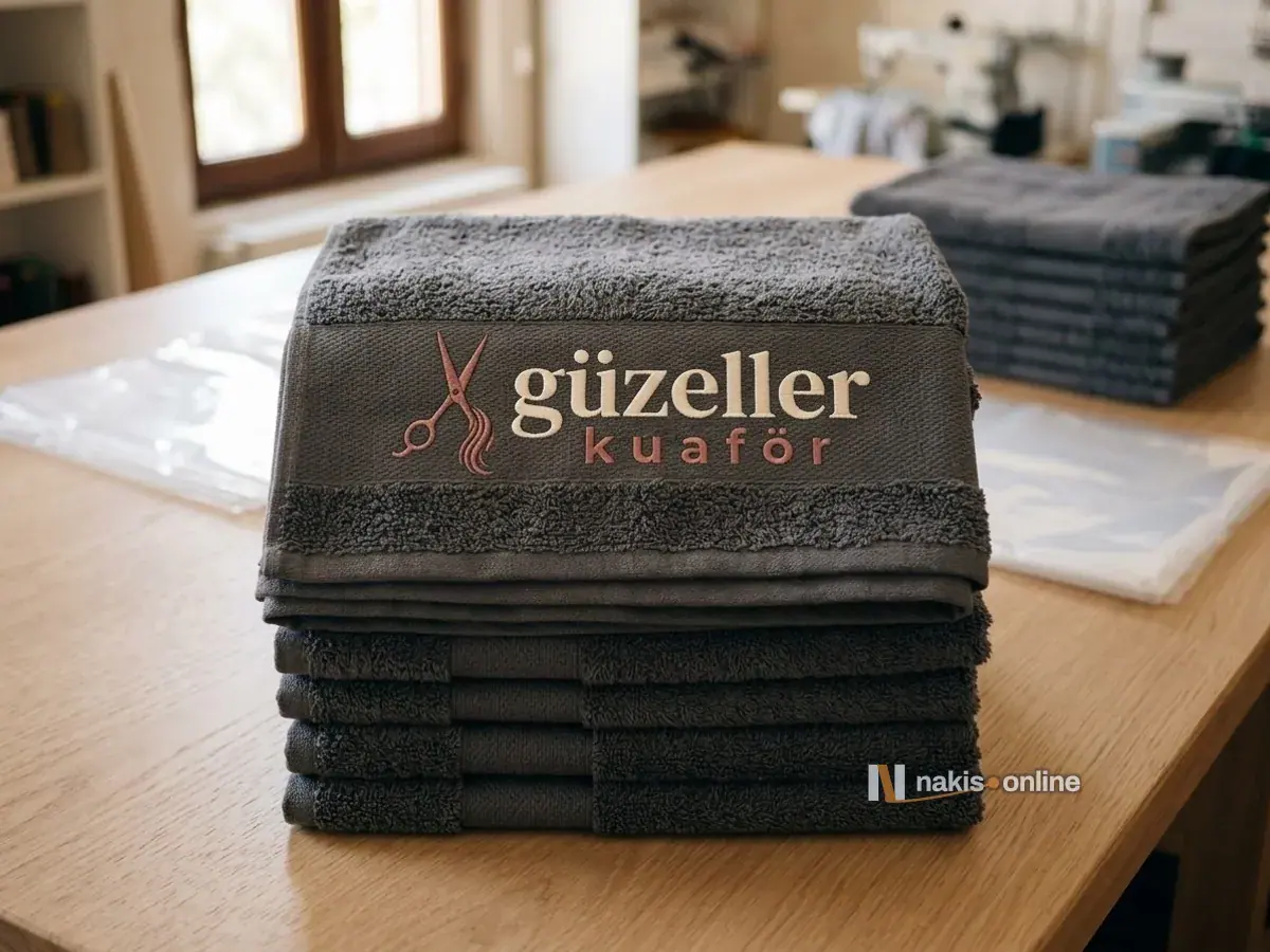 Paketleme masasında katlanmış antrasit el havlusu destesi; en üstteki havlunun bordür bandında gül kurusu makas simgeli, krem serif harfli bir kuaför salonunun logosu, arkada bulanık ikinci deste ve şeffaf poşetler