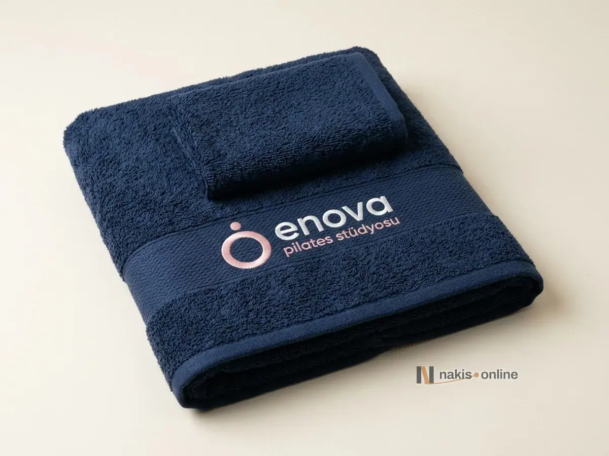 Stüdyo zemininde katlanmış lacivert banyo ve el havlusu; banyo havlusunun dokuma bordür bandında pudra pembesi halka simgeli, beyaz küçük harfli bir pilates stüdyosu logosu
