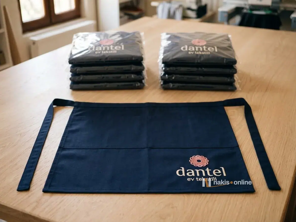 Paketleme masasında önde düz serilmiş lacivert bel önlüğü, arkada şeffaf poşetlere konmuş iki deste önlük; serili önlüğün alt köşesinde pembe rozet simgeli bir ev tekstili mağazasının logosu