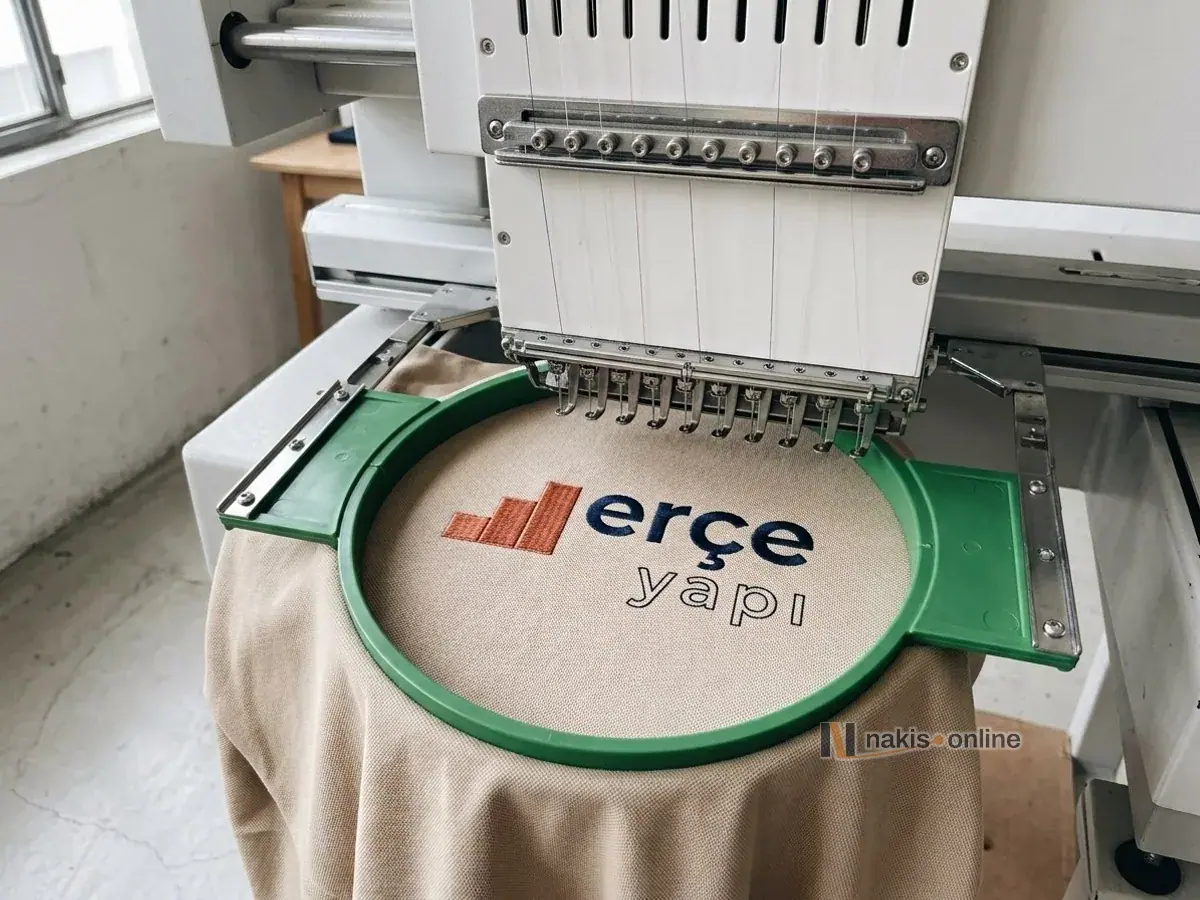 Nakış makinesinin kafası altında yeşil tüp kasnağa gerilmiş kum rengi polo tişörtün sol göğsü; kiremit renkli basamak simgeli bir yapı firmasının logosu yarısı işlenmiş