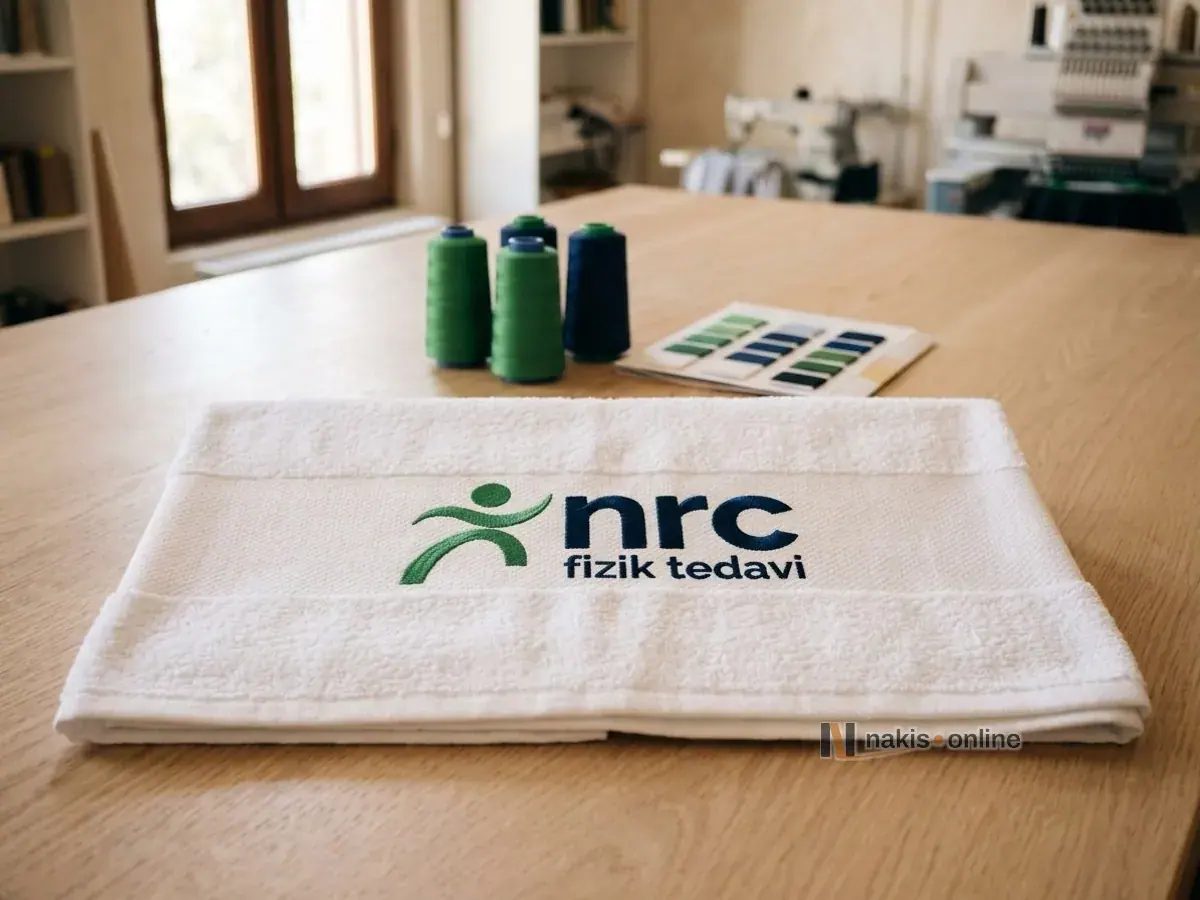 A large white pool towel folded on a light oak table; a physiotherapy centre logo with a green figure and navy lettering is embroidered in the middle of the woven border, with green and navy thread cones and an unlabelled thread colour card behind