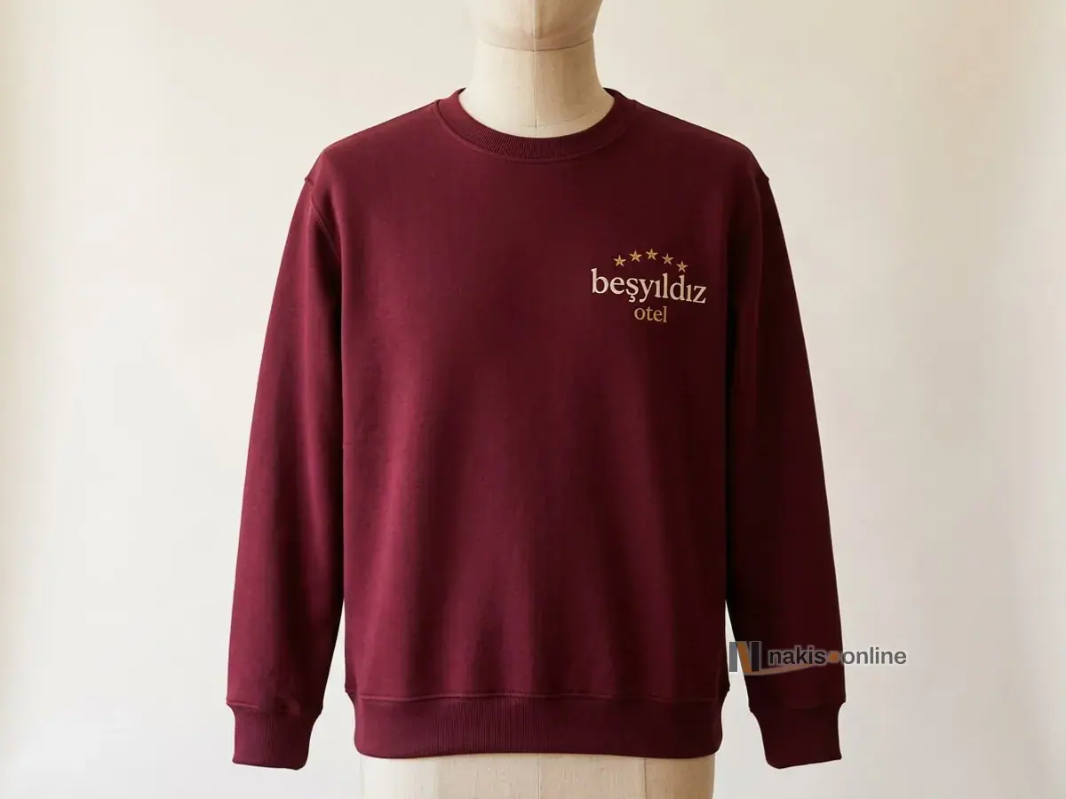 Bordo bisiklet yaka sweatshirt: sol göğüste kemer biçiminde dizilmiş altın sarısı küçük yıldızlar ve krem harflerle bir otelin logosu