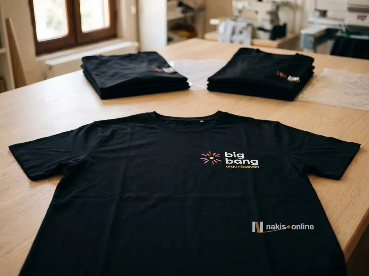 Uzun ahşap masaya düz serilmiş kısa kollu siyah bisiklet yaka tişört; sol göğüste fuşya ve sarı ışınlı simgeyle beyaz harfli bir etkinlik şirketi logosu, arkada şeffaf poşetlerin yanında katlı tişört yığınları, en arkada bulanık dikiş ve nakış makineleri
