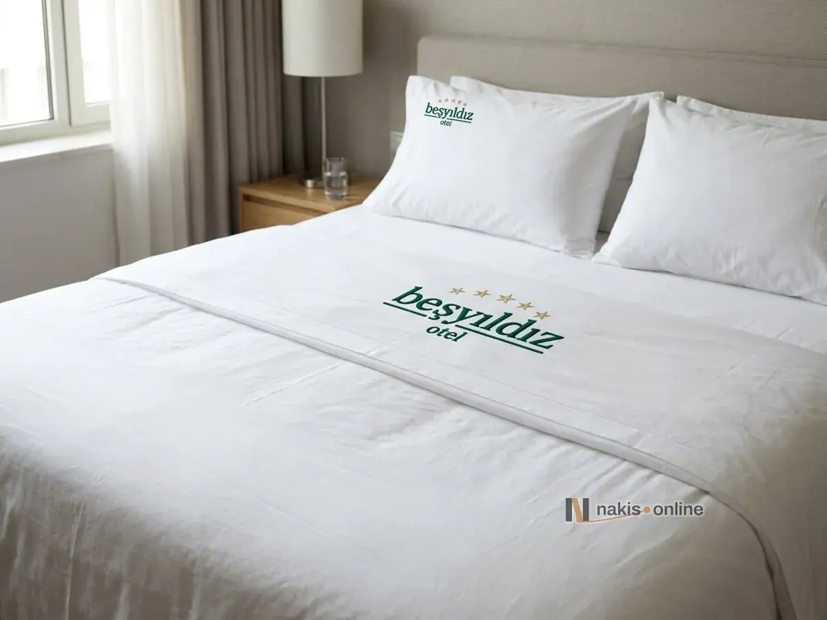 Gün ışığı alan otel odasında beyaz perkal takımla yapılmış çift kişilik yatak; yastık kılıfının köşesinde ve geri katlanan üst çarşafta altın yıldızlı, zümrüt yeşili harfli bir otel monogramı